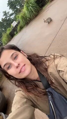 This is a selfie-style video taken from a low angle, showing a young woman in the foreground and a small, furry animal, possibly a rabbit, in the background on a paved pathway. The woman has long, dark, wavy hair and striking blue-green eyes. She is smiling directly at the camera and wearing a tan jacket with a black bag slung across her body. The animal is small and greyish-brown, captured mid-motion on the wet pavement.

The setting appears to be outdoors, with lush green foliage and trees lining the pathway on the left side. A dark stone wall separates the pathway from the greenery. A white sign with red lettering is visible on the left side of the pathway, though the text is too small to be read clearly. The pathway itself is wet, suggesting it may be raining or has recently rained. The lighting is overcast, indicating it could be daytime, possibly morning or afternoon, under cloudy skies. The overall mood of the video seems pleasant, with the woman's smile conveying a sense of joy or amusement at encountering the animal. The location, identified as Seogwipo-si, South Korea, suggests a scenic, possibly natural or park-like environment.