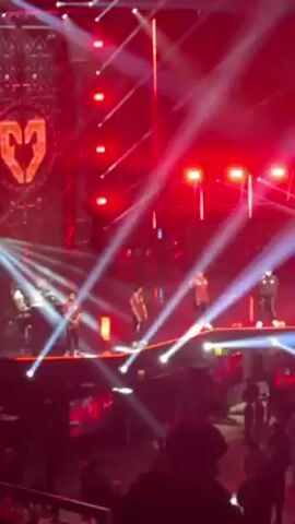 The video shows a group of people on a stage. The stage is lit with red and white lights, including spotlights and neon lights. The backdrop behind the group features a large symbol in red. The people on stage are wearing casual clothing, with some in red shirts. A camera crew is visible to the right of the group. The audience is visible in the foreground, with heads and shoulders seen in the darkness. The stage is elevated, and the overall atmosphere suggests a concert or performance.