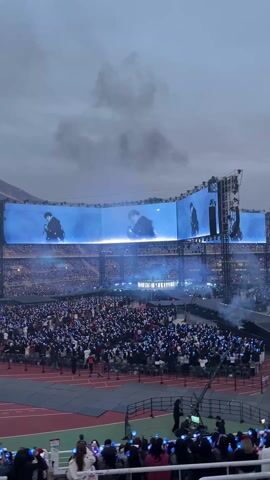 Here's a concise description of the video:

The video shows a massive crowd at a stadium concert, likely a K-pop event. The audience is holding up light sticks, creating a sea of glowing points. A giant screen displays a singer, with the image split across multiple panels. Smoke effects add to the atmosphere. The stadium is packed with people, and the overall scene is vibrant and energetic. The sky is overcast.