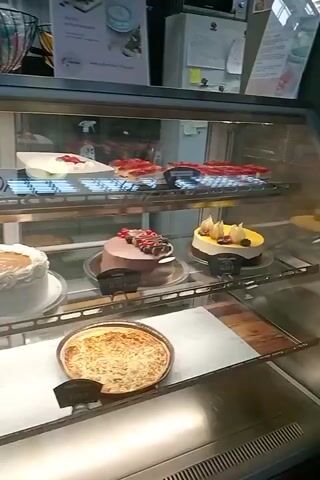 The video shows a bakery display case filled with various baked goods. In the top shelf, there are several white cakes with fruit toppings, as well as what appears to be a few tarts. The middle shelf displays a round white cake with a caramel-colored topping, a chocolate cake adorned with berries, and a small yellow cake topped with fruit. The bottom shelf showcases two large circular pies, one appearing to be a fruit pie. To the left of the display case, there is a counter with a black register and various packaged baked goods. Further back, several people are visible, including a woman in a black shirt and two other individuals blurred in the background, suggesting a busy bakery environment. The overall ambiance appears to be that of a well-stocked and operational bakery.