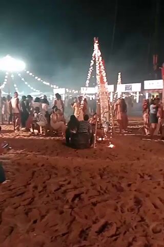 The video shows an outdoor night event on a sandy ground. Numerous people are present, some sitting on the ground or low structures, others standing and mingling. Several tall, pyramid-like structures adorned with string lights are visible, with small fires burning at their bases. In the background, there are various stalls or tents with illuminated signs. A person in a red shirt walks from the left across the frame towards the right. The overall atmosphere appears social and festive.
