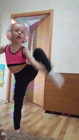 A young girl, with blonde hair tied back in a ponytail and wearing a pink sports bra, black leggings, and headphones with light-up cat ears, is performing a dance or exercise routine in what appears to be a child's bedroom. The girl raises one leg high in the air, then pivots and kicks the leg out to the side. She stumbles a bit, regains her balance, and then spreads her arms as if dancing or balancing. The room has wooden laminate flooring and light-colored walls. A colorful poster is visible on one wall. There's a partially open door with a light visible from the room beyond, as well as a wooden dresser or cabinet.