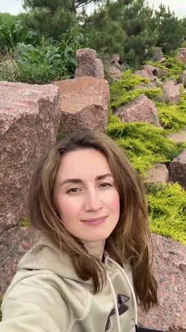 A woman stands smiling in front of a rocky landscape with green vegetation. She is wearing a light beige hoodie with a dark logo on the front. Her brown hair is long and blowing in the wind. The sky above is overcast. The camera angle shifts slightly upwards during the video.