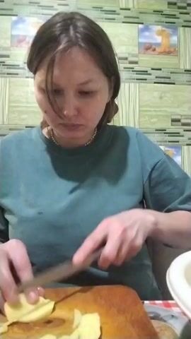 A woman in a teal t-shirt is cutting slices of what appears to be a yellow fruit on a wooden cutting board. She is using a kitchen knife and her hands are in focus. The background is a kitchen wall with decorative tiles.