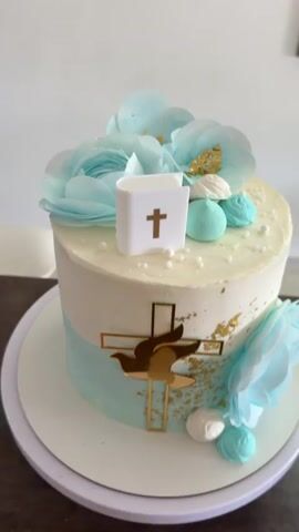 The video shows a close-up view of a decorated cake, likely for a religious celebration like a baptism or first communion. The cake is cylindrical and sits on a white cake stand. The bottom half is light blue, and the top half is white with a slightly textured buttercream frosting. A gold-colored cross with a dove silhouette is affixed to the front. Gold leaf is sprinkled around the cross. The top of the cake is adorned with light blue fabric flowers, small white and blue meringues, and tiny white sprinkles. A small white fondant bible with a gold cross is positioned on top. The background is a plain white wall and floor. The camera slowly rotates, showcasing all sides of the cake. There are some indiscernible sounds in the background, potentially of people talking or playing.