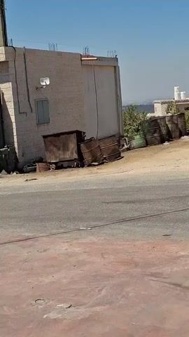 The video pans across a street scene with buildings, vehicles, and barrels. In the foreground, a reddish-pink paved area transitions to an asphalt road. To the left, a partially constructed building with exposed rebar stands next to several rusty metal barrels and a dumpster. Further down the road, more barrels, some green, are lined up. Beyond the barrels, several cars are visible. One car has its hood open, revealing the engine. A silver SUV is parked to the right, followed by another vehicle. A food stand with images of food items is visible near a tree. Finally, a white van with red and gold accents drives by, displaying Arabic text and a coffee logo. The sky is clear and blue.