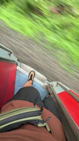 A first-person perspective video shows a person sitting on the edge of a moving train. Their legs are crossed, and they are wearing black shorts and brown and green shoes. A brown and green crossbody bag is on their lap. The train's interior is visible with red and blue seats. Outside, blurred green foliage rushes past, with a dirt track and rocks visible beside the train tracks. The sky is cloudy.