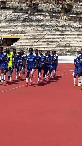 A group of young men in blue soccer uniforms are jogging on a red track in a stadium. The stadium appears to be old and has empty concrete bleachers. Some of the men are wearing yellow goalkeeper gloves. The sky is clear and the sun is shining. The men appear to be warming up or practicing.