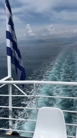 A Greek flag flutters from a white pole on the left side of the frame. The camera is positioned on the deck of a ferry, looking out at the sea. White seats are visible in the foreground. The ferry is moving, creating a wake in the deep blue water. In the distance, mountains are silhouetted against a cloudy sky. The weather appears to be fair with some scattered clouds.