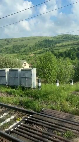 The video shows a view from a moving train. The train tracks are visible in the foreground. The landscape outside the train window includes green fields, trees, and rolling hills under a blue sky with scattered clouds. Houses and small buildings are interspersed in the landscape. The overall environment appears rural and peaceful.