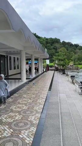 A person walks on a paved walkway next to a river. Another person can be seen walking in the distance near a building. The walkway has decorative white railings with planters. The river is flowing and the surrounding environment is lush with trees. Buildings are visible along the riverbank. The sky is cloudy.