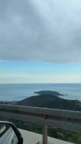 The video shows a view from a moving vehicle, looking out over a body of water and a coastline. In the foreground, the side mirror of a car is visible on the left. A metal guardrail runs along the edge of the road. In the middle ground, there are green, tree-covered hills and a small island with a town or village nestled on the coast. The background consists of a vast expanse of the ocean meeting a partly cloudy sky. The lighting suggests it is either early morning or late afternoon. There are no people visible in the frame.