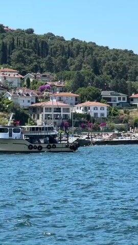 The video captures a serene seascape with a backdrop of lush green hills dotted with houses. The foreground is dominated by the glistening blue water of the sea, rippled by a gentle breeze. A beige-colored boat, identified by text on its side as "KUZEY AKDENİZ," cruises across the frame from left to right. It appears to be a passenger boat or ferry, with several windows visible along its side. Moored near the shore is a white lighthouse with a green stripe near its top, standing on a concrete pier. Behind the lighthouse, several other smaller boats are docked. The midground showcases a picturesque hillside covered in trees and various residences, ranging from multi-story houses with white and red roofs to smaller dwellings nestled amidst the foliage. The overall impression is one of a tranquil, sun-drenched coastal setting, likely a vacation destination. The scene is viewed from another vessel, as indicated by a slight obstruction at the very bottom edge of the frame which appears to be part of a railing or structure on the viewing boat.