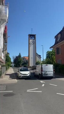 The video shows a street scene with a church tower in the background on a sunny day. The tower is white with a clock face and a cross on top. A white car and a white van drive on the street. Scaffolding is visible on a building to the left. Trees and houses line the sides of the street. Church bells can be heard ringing.