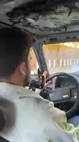 A young man is driving a car with a badly damaged interior. The headliner is torn and hanging down. The driver is wearing a light-colored shirt with yellow and green accents and appears to be recording himself on his phone, which is mounted on the steering wheel. There are other cars visible outside the driver's side window. Music is playing in the background.