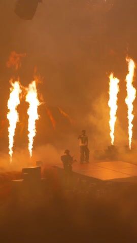 An on-stage performance is shown.  A male entertainer, wearing a white tank top and camouflage pants, stands on a stage as he raps into a microphone.  To his left, a man is playing an electric guitar, and another man films the entertainer with a camera. Behind them are stage decorations resembling a lava flow, and a tower with lights and scaffolding. Flames erupt periodically behind the entertainer, and smoke fills the stage. A crowd is seen in front of the stage.