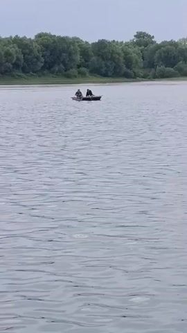 Two individuals are in a small boat on a body of water. The boat is moving forward through the water. A line of trees is visible in the background along the shoreline. The sky appears overcast.