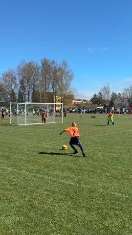 This video captures an outdoor youth soccer match in Ragana, Latvia, on a bright, sunny day with a clear blue sky. The game is being played on a well-maintained green artificial grass field. In the foreground, a child wearing an orange jersey with the number 21 on the back is seen dribbling a yellow soccer ball with their right foot, moving towards the goal.

The main action is centered around the white soccer goal, where several other players are positioned. A goalkeeper, dressed in a black and orange long-sleeved jersey, stands ready to defend the net. Other children in orange jerseys and some in yellow jerseys are visible on the field, actively participating in the game or observing the play.

In the background, beyond the soccer field, there are numerous trees, some with bare branches and others with early spring foliage. A large, light-colored building, possibly a school or community center, can be seen behind the trees. A crowd of spectators, including adults and other children, is gathered along the sidelines of the field. The overall scene depicts a lively and energetic youth sporting event under pleasant weather conditions. No specific text is clearly visible on jerseys or any signs within the frame. The audio includes voices, likely coaches or parents, giving instructions and cheering, with phrases such as "Dod Dušam!" (Give to Dušam!) audible.