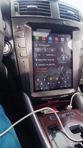 Here is a detailed description of the video:

The video shows the interior of a car, focusing on the dashboard and steering wheel. A large touchscreen display is prominent in the center console, showing a music player interface with songs listed. A smartphone charging cable is plugged into a USB port nearby. The steering wheel is black and has several buttons.

The video then shifts to a first-person view from the driver's seat. A hand wearing a gold Apple Watch with a blue band is seen gripping the steering wheel. Through the windshield, a road stretches into the distance, bordered by open fields with scattered trees. The sky is overcast. The perspective implies the car is in motion.