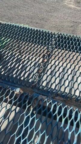 The video begins with a close-up of a metal grate, then pans up to reveal a metal bucket on a green surface. The scene appears to be an outdoor industrial or construction site with gravel on the ground. In the background, there are trees, buildings, and construction equipment such as a crane and trucks. The sun is shining brightly in a blue sky with some clouds. The camera continues to pan upwards, showing more of the sky and trees. The video ends with a view of a partially open door in the foreground, with the bright sun visible through the opening.