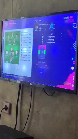 A television screen displays a soccer video game. The screen is split into two halves, showing different team lineups and player information. The left side of the screen shows a formation labeled "Bayern München" with player portraits and names. The right side of the screen shows a formation labeled "Chelsea" with player portraits and names, as well as a player's individual stats labeled "Tyrigue George". The game appears to be paused or in a menu screen. The television is mounted on a wall with visible cables connected to it. The wall itself has a textured, possibly concrete or stucco, surface. An electrical outlet is visible on the wall to the left of the television.