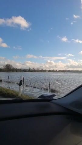 A car interior view shows an expansive flooded field under a bright blue sky with scattered white clouds. The water stretches far into the distance, obscuring the ground except for fence posts and shrubbery emerging from the water. In the distance, a small town is visible, marked by trees and a church steeple. The camera is stationary, presumably filming from inside a parked car. The dashboard is visible in the foreground, creating a sense of being inside the vehicle.