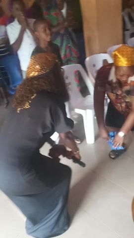 This video captures an indoor gathering, likely a religious or spiritual event, in Nkwelle Ezunaka, Nigeria.

In the foreground, a woman wearing a black abaya or dress with a gold design on the back and a gold-colored curly wig or head covering is kneeling or bowing. Directly in front of her, another woman with a yellow headscarf and a patterned top is seated on a white plastic chair. She is holding a small blue cloth and appears to be engaged in an action with her hands, possibly wiping or blessing something on the floor or near the kneeling woman.

In the background, several other people, including adults and children, are present. Some are seated on white plastic chairs, while others stand. Many are observing the activity in the foreground, with some individuals visible with their hands raised, suggesting participation in worship or prayer. A golden cymbal and part of a drum set are visible behind the seated woman, indicating the presence of musical accompaniment.

The setting appears to be an informal gathering space with a tiled floor and light-colored walls. The overall atmosphere seems reverent and possibly emotional, accompanied by what sounds like gospel or praise music playing in the background. No discernible text is visible on clothing or in the surroundings.