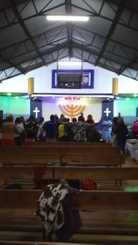 In an indoor, medium-shot video, a group of people are gathered inside a building, likely a church, and are actively dancing and moving. The building has a metal roof structure and rows of wooden pews. The people are dressed in a variety of clothing, and their movements suggest they are engaging in some kind of religious ceremony or celebration. At the front of the room, a stage is decorated with crosses and a menorah-like symbol, illuminated by various colored lights. A screen is also visible on the stage. In the foreground, some clothing items are draped over the pews.