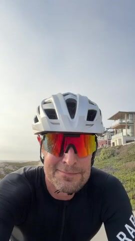 A man wearing a white helmet and red-tinted sunglasses is riding a bicycle on a paved path. He is wearing a black long-sleeved cycling jersey. The path is next to a sandy beach with some vegetation. In the background, there is the ocean and a modern house on a hill. The video briefly pans to show more of the beach and other people riding bicycles in the distance. The man looks directly at the camera and smiles.