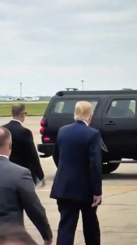 The video shows a group of men, presumably security personnel, escorting a man with blonde hair in a dark suit towards a large black vehicle. The man in the blonde hair appears to be the person of interest. The environment appears to be an outdoor tarmac with a cloudy sky. One security guard in the foreground is wearing sunglasses and a dark suit. The other security guards are wearing dark suits and are positioned around the man in the blonde hair. The black vehicle is a large SUV or similar vehicle, and the man in the blonde hair is walking towards the rear passenger door. The video is shaky and the focus is not consistently sharp, suggesting it was filmed from a distance or by someone moving.