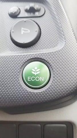 The video shows a close-up of a car's interior, focusing on a green button labeled "ECON" with a leaf icon above it. The button is circular and appears to be made of plastic. It is set into a textured gray dashboard panel. Above the ECON button, there are two other circular buttons, one with a symbol that looks like a car door or trunk, and another with "L R" which likely indicates left and right mirror adjustment. Below the ECON button, a portion of other buttons or controls are visible, but they are out of focus. The lighting suggests the video was taken inside a car during the daytime, with some ambient light creating subtle reflections on the dashboard. There are no people visible in the video. The camera is held steady, with slight movements between frames, likely due to hand holding the camera. The primary action is the static display of the ECON button, with slight variations in focus and angle.