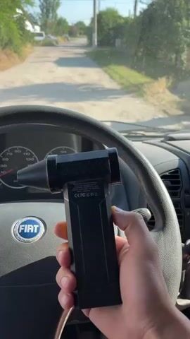 A first-person perspective video shows a person holding a portable anemometer in their right hand. The anemometer is black and yellow and has a spinning impeller at the front. The person is sitting in the driver's seat of a Fiat van. The steering wheel is visible, as is the dashboard with the speedometer and tachometer. The van is driving on an unpaved road, with vegetation on either side. The sky is clear and the sun is shining. The video appears to be filmed during the day. The person briefly moves their left hand to the steering wheel.