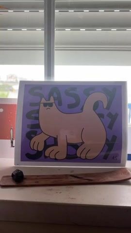 The video shows a framed picture of a tan cat wearing sunglasses and the word "SASSY" written in large black letters repeatedly behind the cat against a purple background. The picture is placed on a windowsill in front of a window with white blinds. To the left of the picture is a small, dark, irregular object resting on a wooden incense holder. The camera zooms in slightly on the picture and then zooms back out.