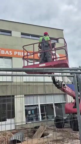 A worker in a red hard hat and gray overalls operates a cherry picker to repair a sign on the side of a building. The building has a large orange and white sign that reads "PRECES" and "alba". A chain-link fence is in the foreground, and a construction site with debris is visible behind it. The sky is overcast.