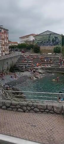 The video shows an outdoor scene with a body of water and a rocky shoreline. The water is a clear, greenish-blue color and several people are swimming or wading in it. Along the shore, there are tiered, rocky steps where many people are sitting, sunbathing, or observing the water. Some individuals are visible on a paved walkway in the foreground, with a metal railing separating them from the scene. Behind the steps, buildings and dense green foliage can be seen, suggesting a coastal or riverside location with some urban development. The sky is overcast, indicating cloudy weather.
