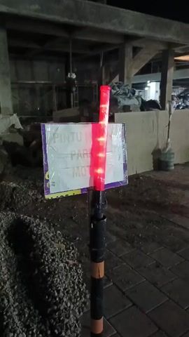 A red flashing traffic wand is positioned in front of a sign that appears to say "PINTU MASUK PARKIR MOTOR" in Indonesian. The wand is mounted on a black pole with orange bands. The sign is covered in clear plastic sheeting, and behind it, there is a dark, somewhat cluttered background with what looks like construction materials and scaffolding. The ground in the foreground is covered in gravel and paving stones. The lighting suggests it is nighttime or dusk.