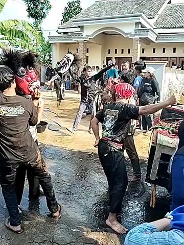 A group of people are gathered in front of a house, which appears to be a home, possibly in a village or rural area, with a yard and trees. The ground is wet, suggesting recent rain or water being present. In the foreground, several individuals are dressed in traditional costumes, some with elaborate masks resembling animals or mythical creatures, indicative of a cultural performance or festival. One person is wearing a red bandana. People are interacting, with some appearing to be performing and others watching. The overall atmosphere seems lively and communal.