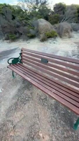The video shows a scenic view from a rocky outcrop overlooking the ocean. The video begins with a close up of a wooden bench with green armrests. The camera pans up to show a landscape of low green shrubbery and a rocky path leading to a cliff edge. The camera continues to pan and the vast blue ocean comes into view under a partly cloudy sky. In the distance, a few islands are visible and a boat cuts through the water leaving a white wake.