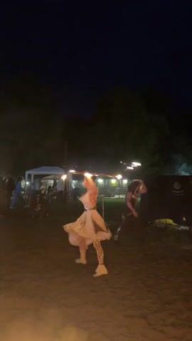 A woman in a white dress dances with flaming hoops at night. She is in a field with trees in the background and people watching. There is also a fire pit in the foreground. Another woman in a dark outfit is visible in the background, also performing.