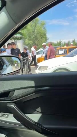 The video captures a scene on a sunny day in Yasnaya Polyana, Russia. The view is from inside a car, looking out the passenger side window. In the foreground, the car's interior is visible, including the door panel and a side mirror reflecting a street scene.

The main focus is an outdoor gathering of people, seemingly involved in a discussion or minor incident. A group of individuals, including several young boys and older men, are standing around. One boy is on a bicycle. Two men appear to be interacting directly, perhaps exchanging words or resolving an issue.

There are several vehicles present: a white car, an orange vehicle, and another white car. The presence of multiple cars and people suggests a public area, possibly a street or parking lot. Trees and a building are visible in the background, under a bright blue sky with scattered clouds, indicating pleasant weather.

The overall atmosphere appears to be calm, though the gathering of people and their focused interaction might suggest a minor dispute or a noteworthy event unfolding. The time of day is clearly daytime, likely late morning or afternoon, given the bright sunlight and shadows. No visible text is present in the video.