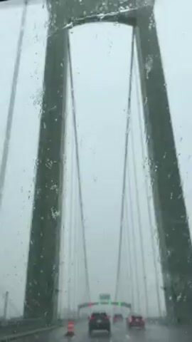 The video shows a car driving on a bridge on a rainy day. The bridge is a suspension bridge, with tall towers and cables visible through the windshield. There are other cars on the bridge, and traffic signs indicate the directions of various routes. The windshield wipers are on, clearing the rain. The sky is overcast and gray, contributing to the low visibility.