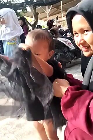 A child in a black dress is being held by an adult wearing a dark hijab and a maroon top. The child is facing away from the camera. Another person in a white hijab and a blue shirt is visible behind them, along with several motorcycles and trees in the background. The ground is paved with bricks. The adult appears to be smiling and holding the child's left arm. The child's right arm is extended forward.