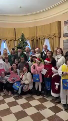 A group of about 25 people, mostly children and adults, stand in front of a Christmas tree and window with gold-colored curtains in a room with a black and white checkered floor. The children are holding blue gift bags. Several adults are dressed in Santa Claus hats or robes. There are two men in clerical garb. Most of the people are looking at the camera, and several are smiling. A man speaks in the background.