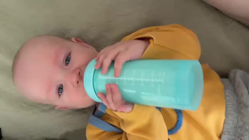 A baby with blue eyes and a yellow shirt is lying down and drinking from a teal baby bottle. The bottle has measurement markings on it. The baby's hands are holding the bottle. The background appears to be a bed or couch with a textured light green surface.