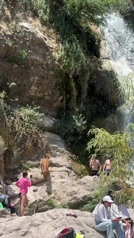 The camera shows people gathered near a waterfall on a sunny day. 
A woman in a pink top and dark bottoms stands on a rock taking a photo. A golden retriever is next to her. Two shirtless men are standing on rocks closer to the base of the waterfall. Several people are seated on the rocks in the foreground. A man in a grey shirt and baseball cap is pointing towards the waterfall. Backpacks and other items are scattered nearby. The area around the waterfall is rocky with lush green vegetation covering the cliff faces.