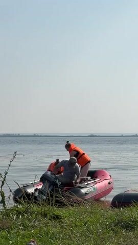 A man and two young women are in an inflatable boat near a grassy shore. The man, wearing a cap and a gray t-shirt, is sitting in the boat and appears to be adjusting something on the engine. One woman, in an orange life vest, is reaching over the side of the boat. The other woman, also in an orange life vest, is sitting in the boat looking at her phone. The boat is partially in the water, and another inflatable boat is visible to the right. The background shows a body of water and a hazy horizon. The overall lighting suggests it is daytime, likely sunny.