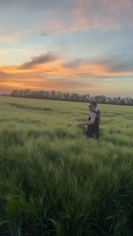 The video shows a man standing in a field of tall green grass with a sunset in the background. The man is wearing a black Chicago Bulls jersey with the number 1 on it, black shorts, and sunglasses. He is facing the camera with his arms outstretched. The sky is filled with orange, pink, and purple clouds. The field is on a slight hill, and there is a line of trees in the distance.