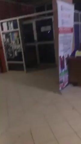 The video appears to be taken in a dimly lit interior space, potentially a hallway or a lobby. The camera moves around, showing different parts of the area. There are walls, columns, and structures that appear to be part of the building's architecture. One section features what looks like a partitioned office area with a white frame and dark panels. In another direction, there's a red wall section. The overall environment appears to be relatively empty, with a focus on the structural elements and the available light, creating a somewhat dark and indistinct atmosphere.