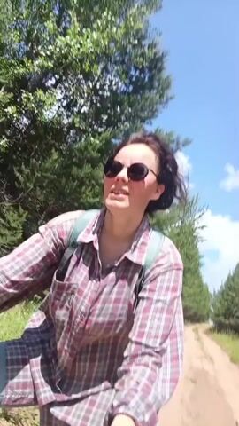 A young woman with dark hair pulled back wears sunglasses and a red and white plaid shirt. She is wearing a backpack and is on a bicycle. She is smiling and looking off to the side. She appears to be on a dirt road surrounded by trees. The sky is blue with some clouds.