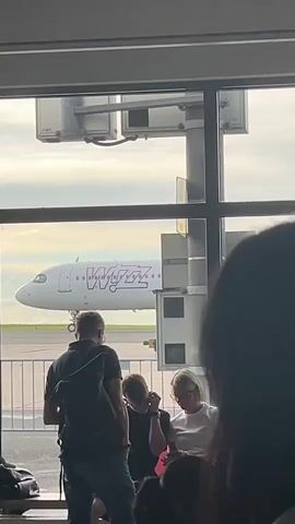The video captures an airport scene, likely at a gate in Dortmund, Germany, with a view of the tarmac through large windows. Inside the terminal, several people are waiting. In the foreground, a man with a backpack stands facing away from the camera, looking towards the window. To his right, two women are seated and conversing. One woman wears a white t-shirt and glasses, while the other is in a dark top. A young man in a red hoodie is also seated further to the left. A black wheeled object, possibly a wheelchair or luggage, is visible in the lower right.

Outside, a white Wizz Air aircraft is parked on the tarmac. The airline's name, "Wizz," is prominently displayed in large purple lettering on the fuselage, and "wizzair.com" is visible below it. The plane features distinctive pink engine nacelles. The sky appears bright but overcast or partly cloudy, indicating daytime. Airport infrastructure, including metal structures and parts of a jet bridge, are visible between the waiting passengers and the aircraft. The overall scene depicts a typical airport waiting area with passengers awaiting their flight.